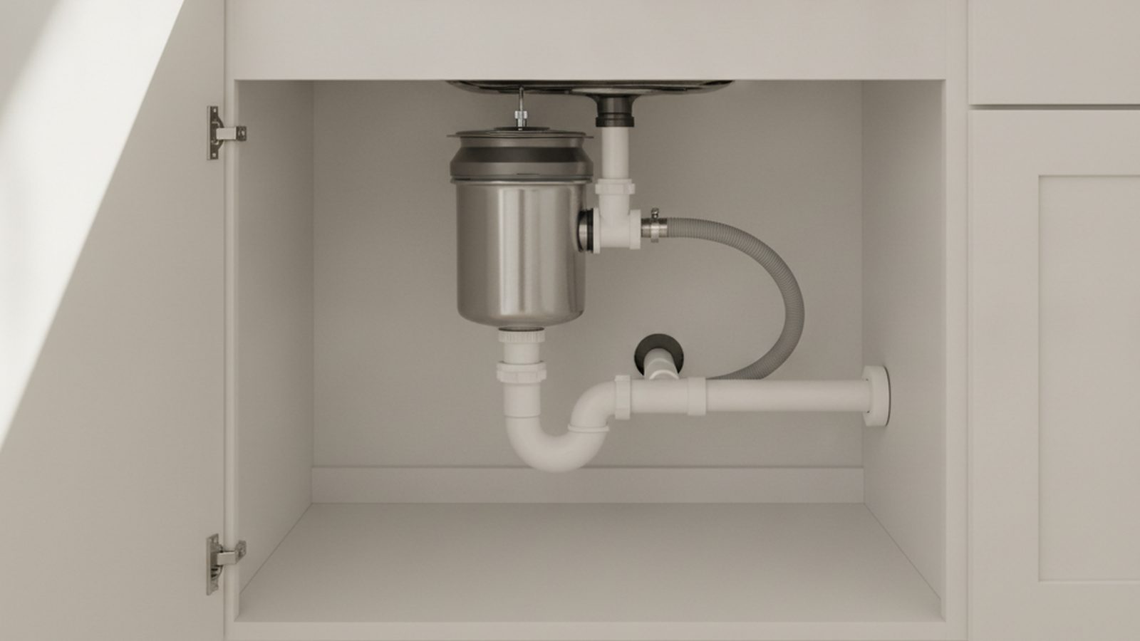 Stainless steel garbage disposal mounted under a kitchen sink with a white PVC drain trap and dishwasher hose